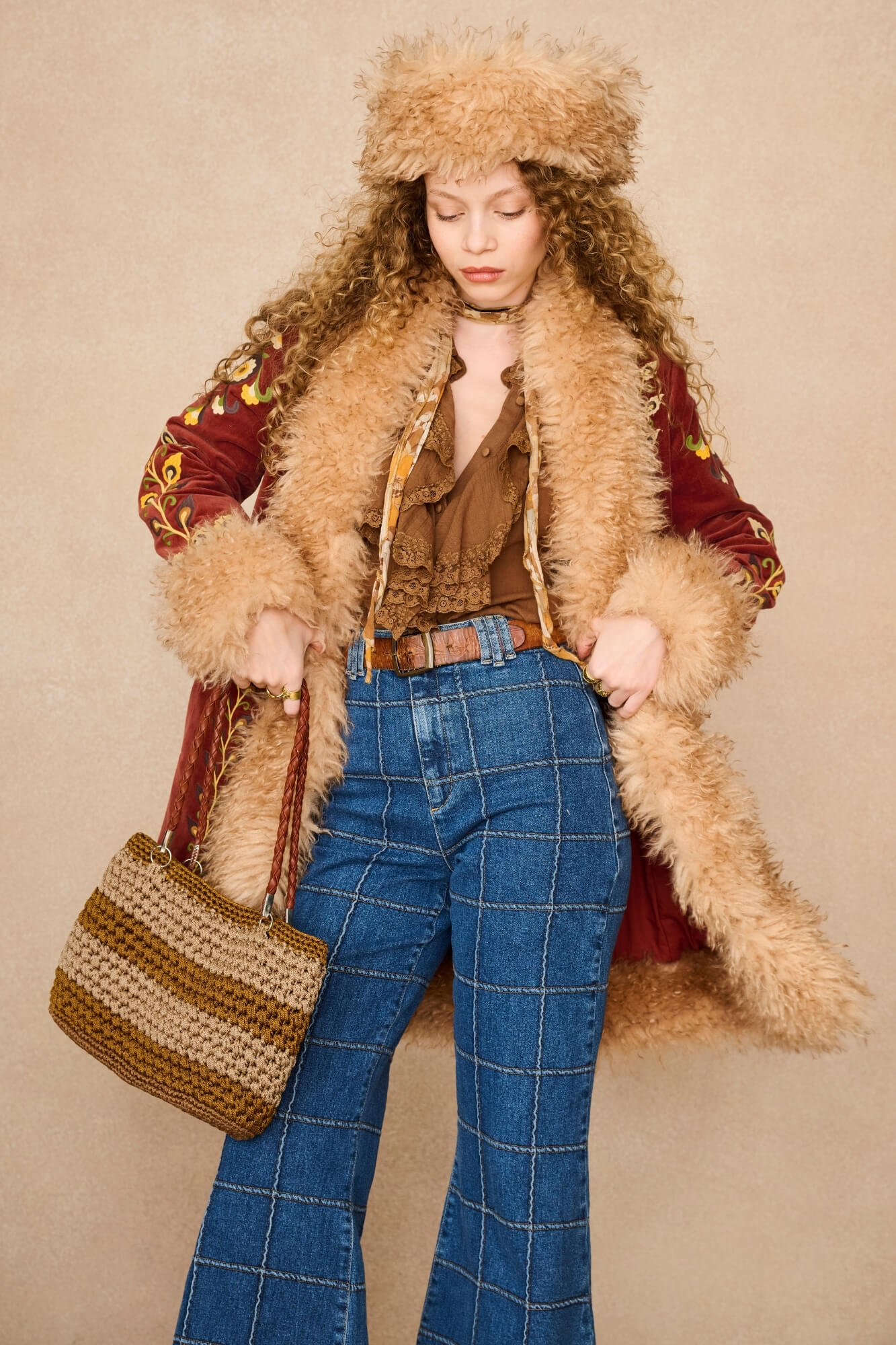 A model wearing a bohemian-style outfit featuring a brown faux fur coat with a matching furry hat, a brown ruffled top, blue plaid flared jeans, and carrying a woven straw bag against a beige background.