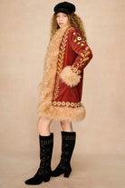 A model wearing a vintage-style rust red velvet Penny Lane coat by The Hippie Shake with beige fur trim, gold floral embroidery, and a black beret, paired with black knee-high studded boots, against a beige studio backdrop.
