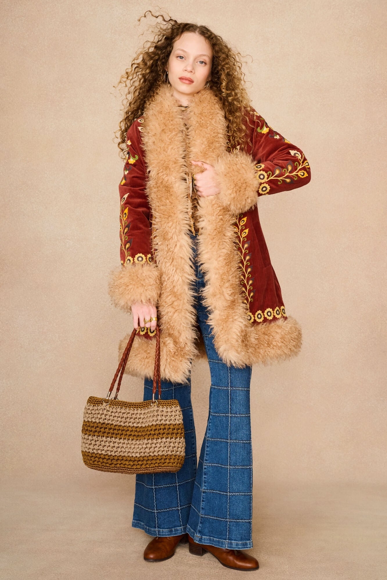 A model wearing a vintage-style red velvet Penny lane by The Hippie Shake coat with beige fur trim and gold embroidery, paired with blue plaid flared jeans, brown boots, and a woven tan handbag, set against a plain beige studio backdrop.