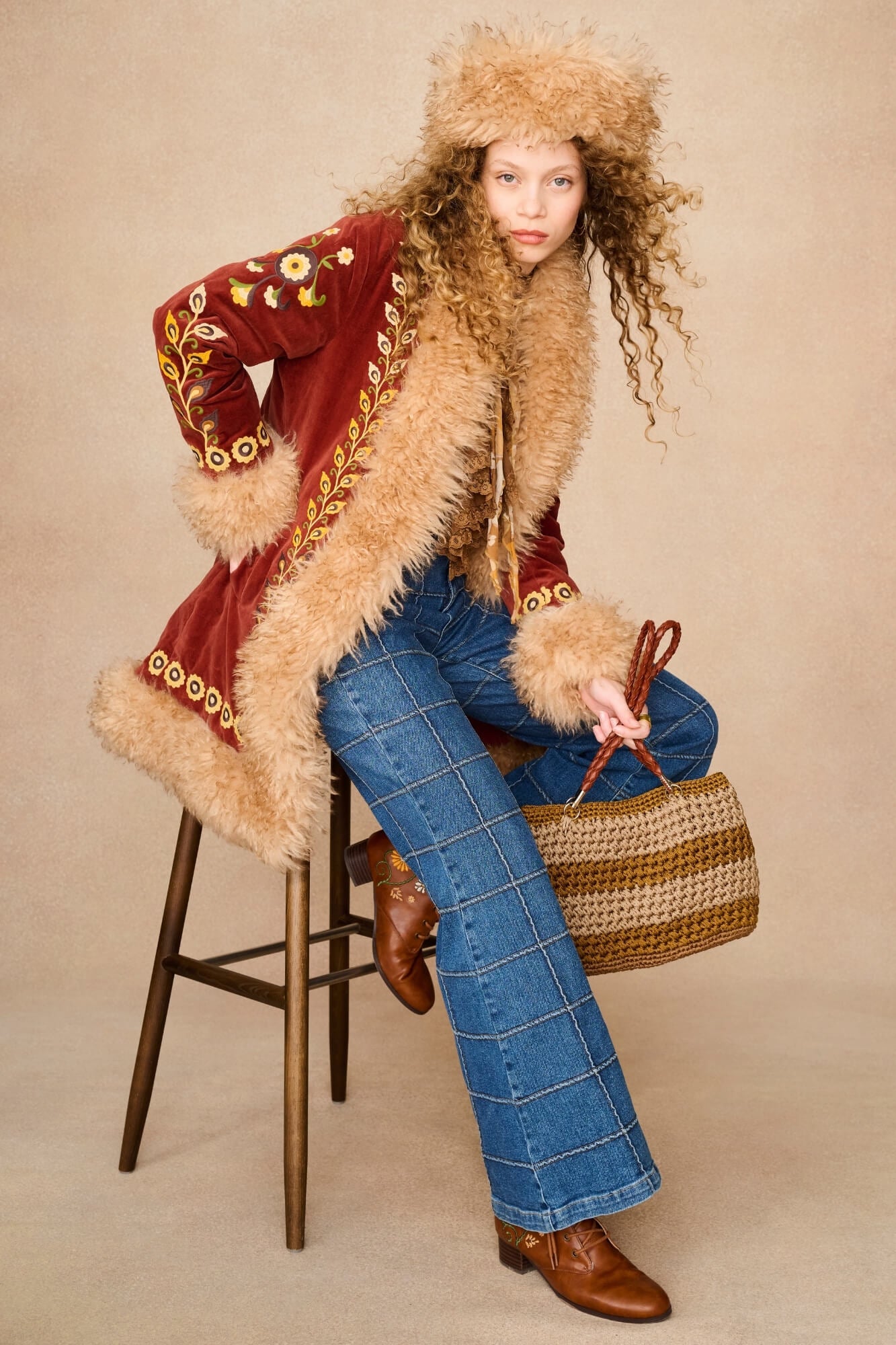 A model sits on a wooden stool wearing a vintage-style red velvet jacket with beige fur trim, a matching fur-trimmed hat, blue plaid jeans, brown leather boots, and holding a woven straw handbag against a beige studio background.
