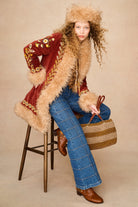A model sits on a wooden stool wearing a vintage-style red velvet jacket with beige fur trim, a matching fur-trimmed hat, blue plaid jeans, brown leather boots, and holding a woven straw handbag against a beige studio background.