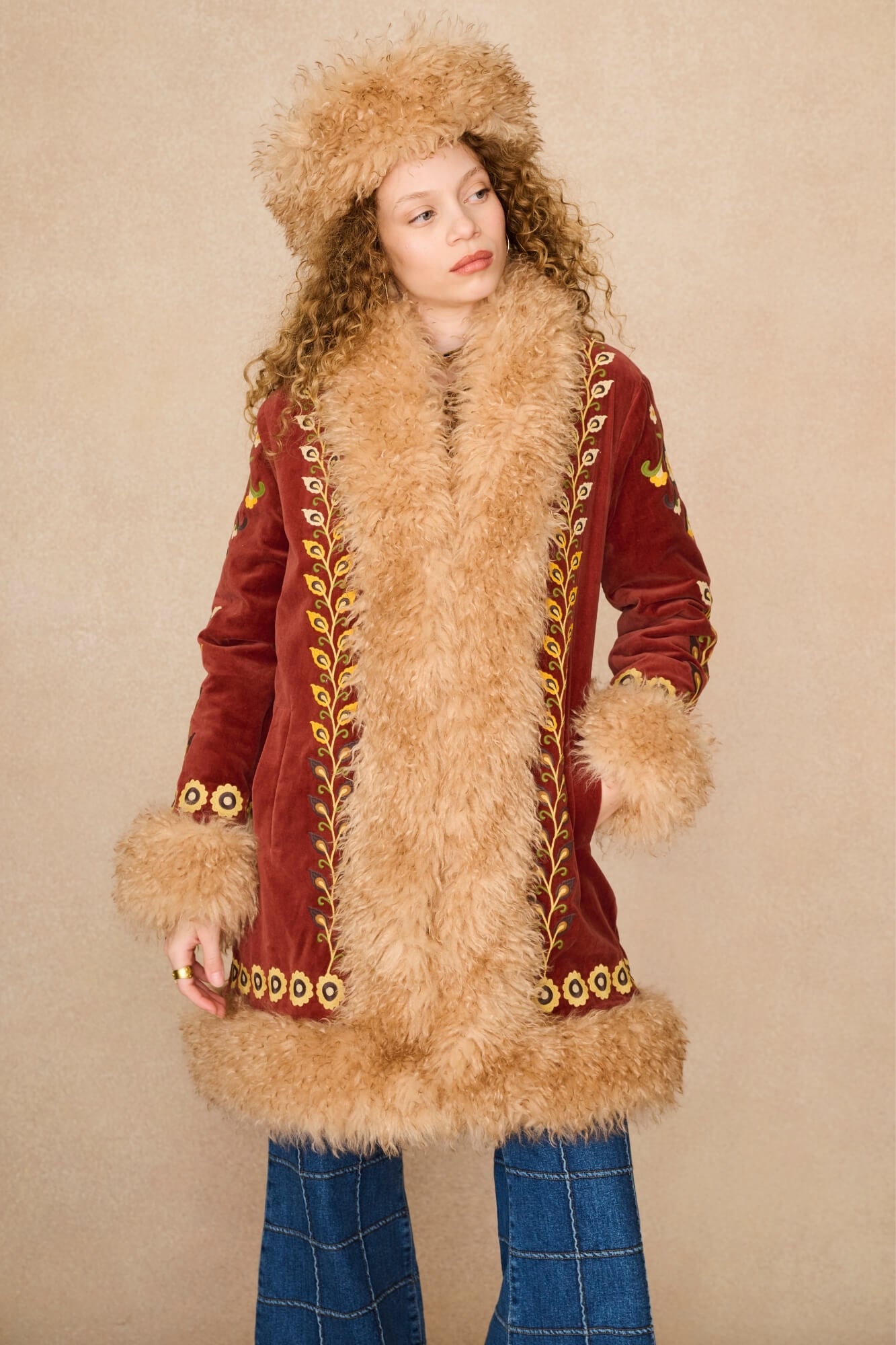 A model wearing a vintage-style mid-length Penny Lane coat by The Hippie Shake made of deep red velvet with a large beige fur collar, cuffs, and hem, decorated with gold embroidered leaf patterns. She is also wearing a matching fur-trimmed hat and blue plaid pants, set against a plain beige background.