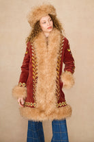 A model wearing a vintage-style mid-length Penny Lane coat by The Hippie Shake made of deep red velvet with a large beige fur collar, cuffs, and hem, decorated with gold embroidered leaf patterns. She is also wearing a matching fur-trimmed hat and blue plaid pants, set against a plain beige background.