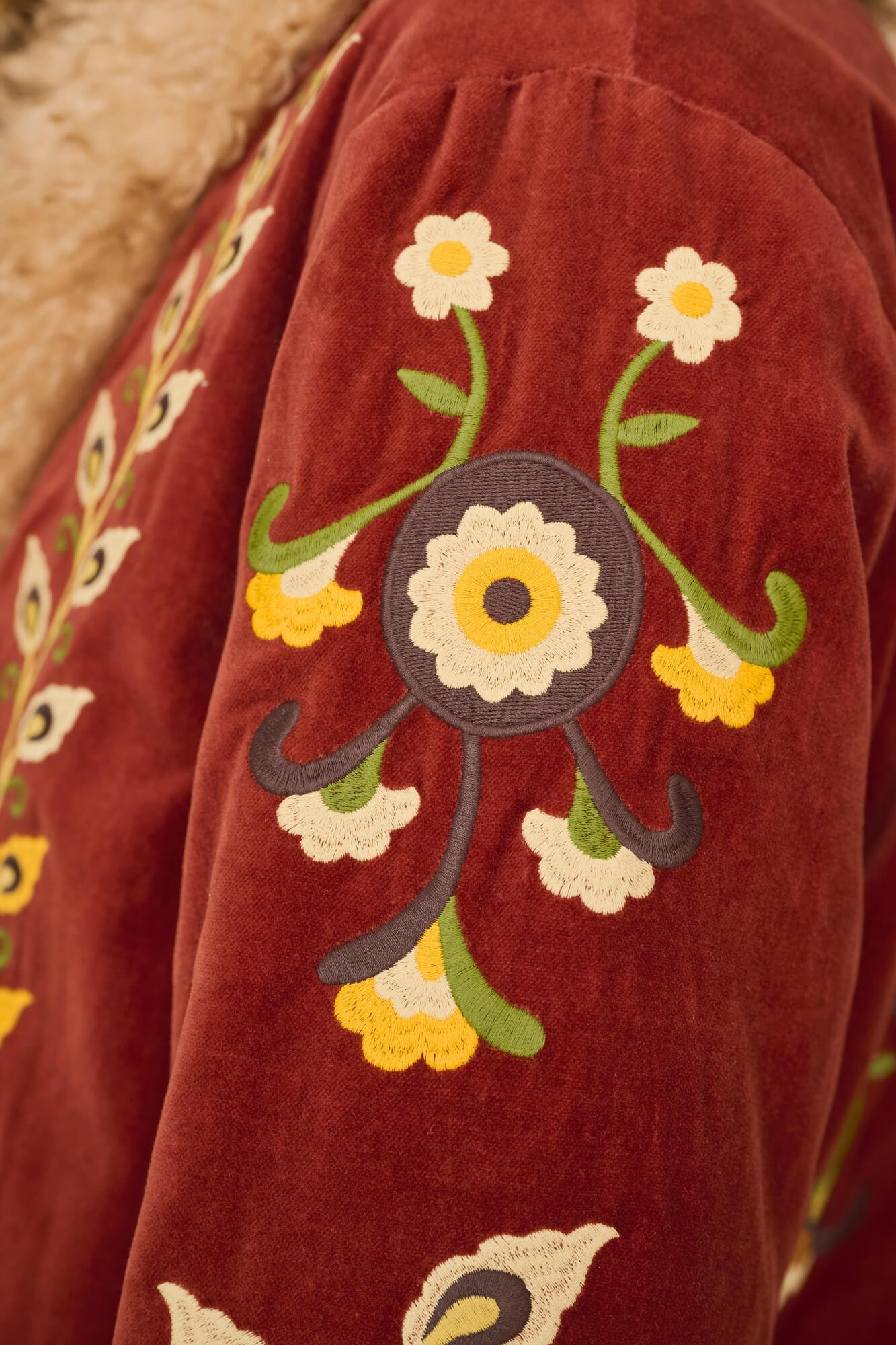 Close-up of a rust red velvet jacket with embroidered floral patterns on the sleeve, featuring white, yellow, and green details.