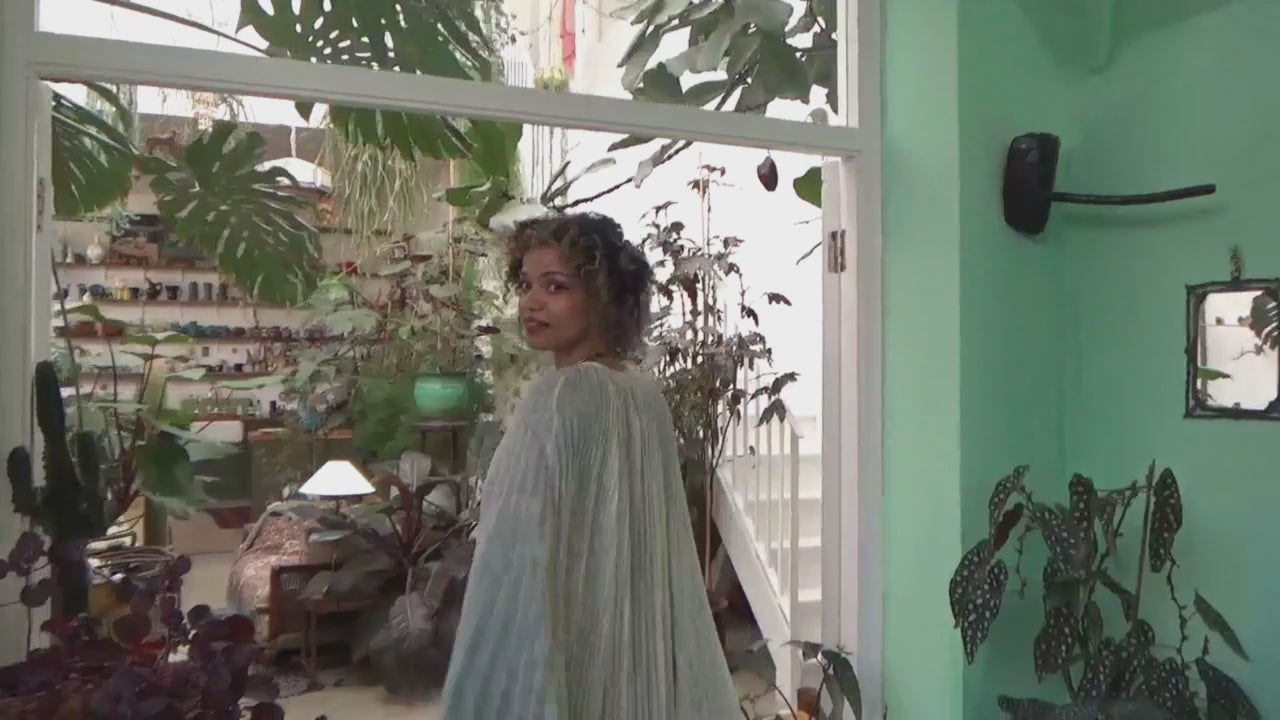 Model in a 70s inspired white cape floating through a greenhouse with pot plants and greenery