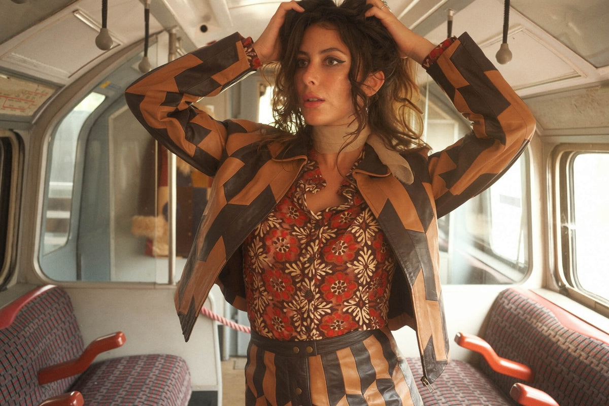 A woman posing inside a vintage london tube train wearing a brown and black geometric patterned jacket over a red and beige floral print top and striped trousers.